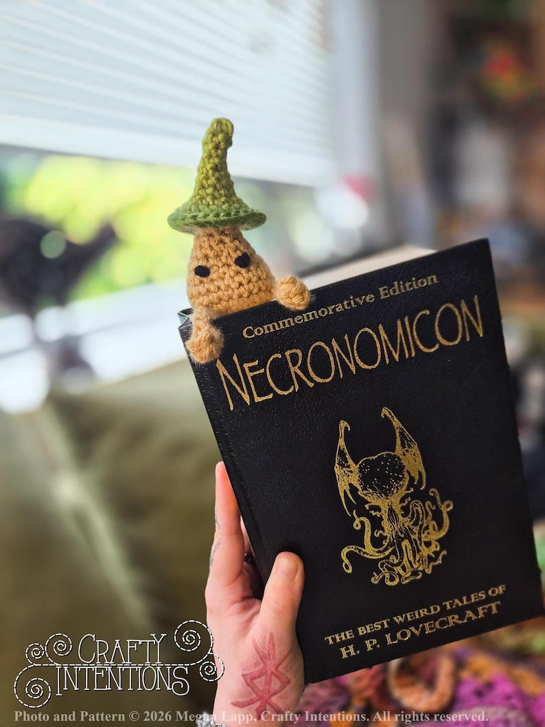 Puede incluir: Un marcap&aacute;ginas de ganchillo con forma de hongo caprichoso con un sombrero verde, asom&aacute;ndose de un libro negro titulado "NECRONOMICON". El libro tiene letras doradas y una ilustraci&oacute;n dorada. Tambi&eacute;n es visible el texto "THE BEST WEIRD TALES OF H. P. LOVECRAFT".