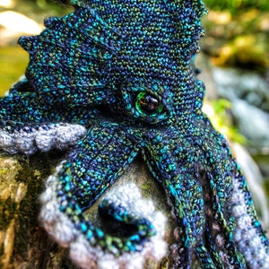 May include: A crocheted octopus amigurumi toy with a green eye, blue, green, and black yarn, and a gray and white yarn for the tentacles. The octopus is sitting on a piece of wood.