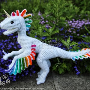 May include: A white crocheted dinosaur with rainbow coloured wings and a horn. The dinosaur is standing on a bed of purple flowers.
