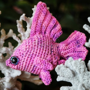 Puede incluir: Un pez betta de crochet rosa con un ojo azul brillante, sentado en un arrecife de coral blanco. El pez es un juguete amigurumi hecho a mano.