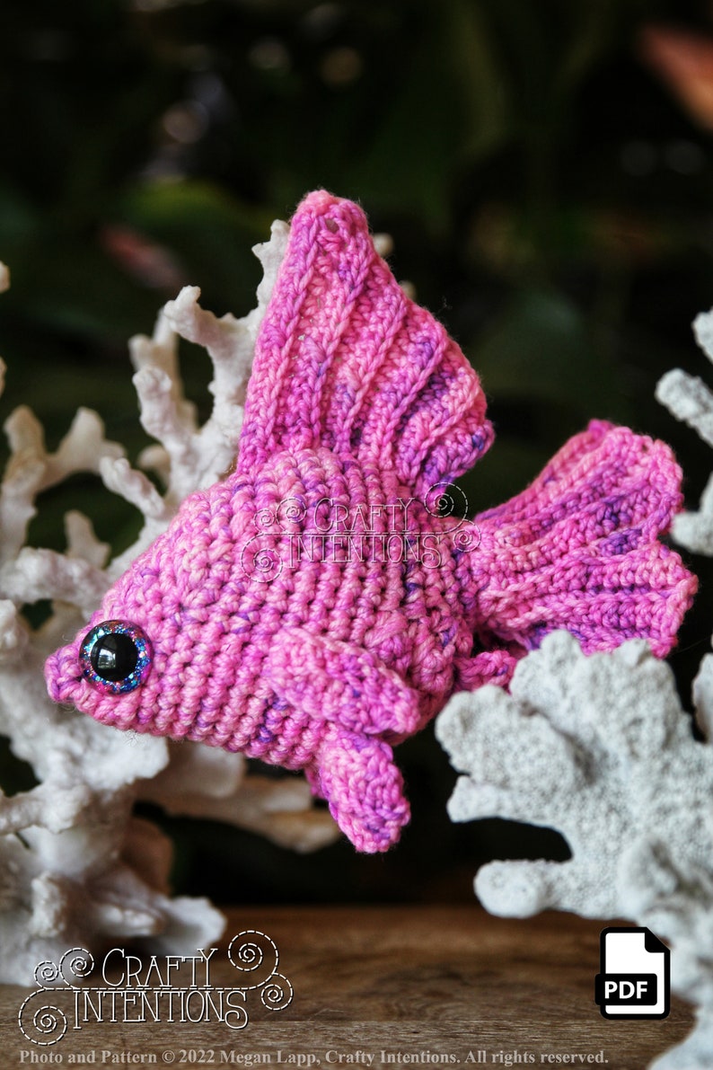Puede incluir: Un pez betta de crochet rosa con un ojo azul brillante, sentado en un arrecife de coral blanco. El pez es un juguete amigurumi hecho a mano.