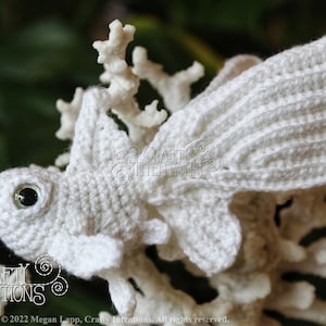 Puede incluir: Pez amigurumi blanco tejido a crochet con un ojo negro, sentado en una rama de coral blanca. El pez tiene una aleta caudal grande y una aleta dorsal m&aacute;s peque&ntilde;a. La imagen incluye el texto "CRAFTY INTENTIONS" y "PDF".