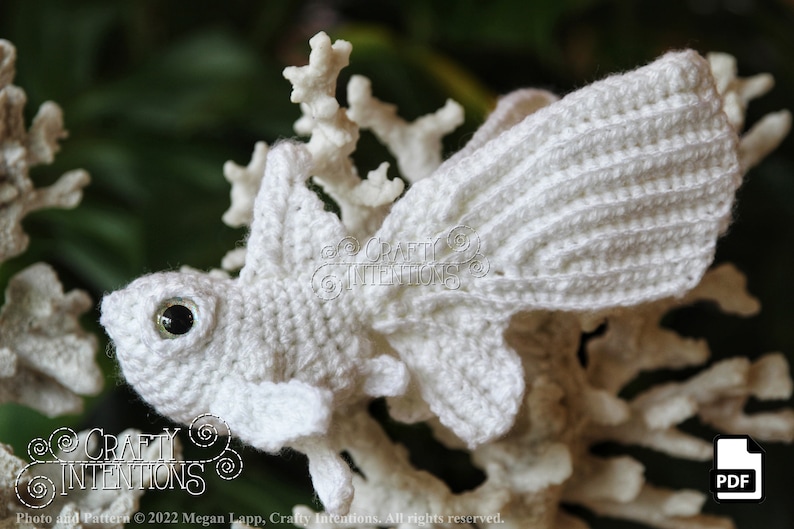 Puede incluir: Pez amigurumi blanco tejido a crochet con un ojo negro, sentado en una rama de coral blanca. El pez tiene una aleta caudal grande y una aleta dorsal m&aacute;s peque&ntilde;a. La imagen incluye el texto "CRAFTY INTENTIONS" y "PDF".