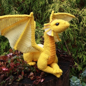 May include: A yellow crocheted dragon with wings outstretched. The dragon is sitting on a tree stump with green foliage in the background.