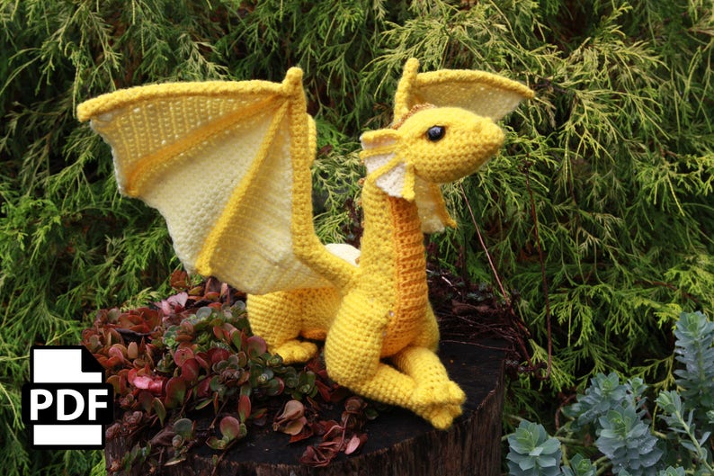 May include: A yellow crocheted dragon with wings outstretched. The dragon is sitting on a tree stump with green foliage in the background.