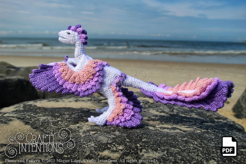 May include: A crocheted dinosaur toy with purple, pink and white feathers. The dinosaur is standing on a rock with the sea in the background.