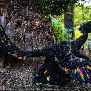 May include: A black crochet dragon with rainbow accents. The dragon is posed with its wings spread and its head tilted back. The dragon is sitting on a tree stump in a woodland setting. The image includes the text "CRAFTY INTENTIONS" and a PDF icon.