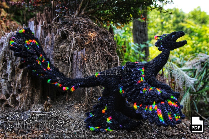 May include: A black crochet dragon with rainbow accents. The dragon is posed with its wings spread and its head tilted back. The dragon is sitting on a tree stump in a woodland setting. The image includes the text "CRAFTY INTENTIONS" and a PDF icon.