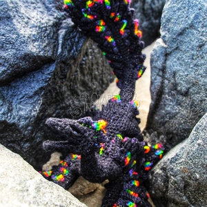 May include: A black crocheted dragon with rainbow coloured accents is posed in a rocky crevice. The dragon is standing on its hind legs with its front legs outstretched. The text "CRAFTY INTENTIONS" is visible in the image. A PDF icon is also visible in the image.