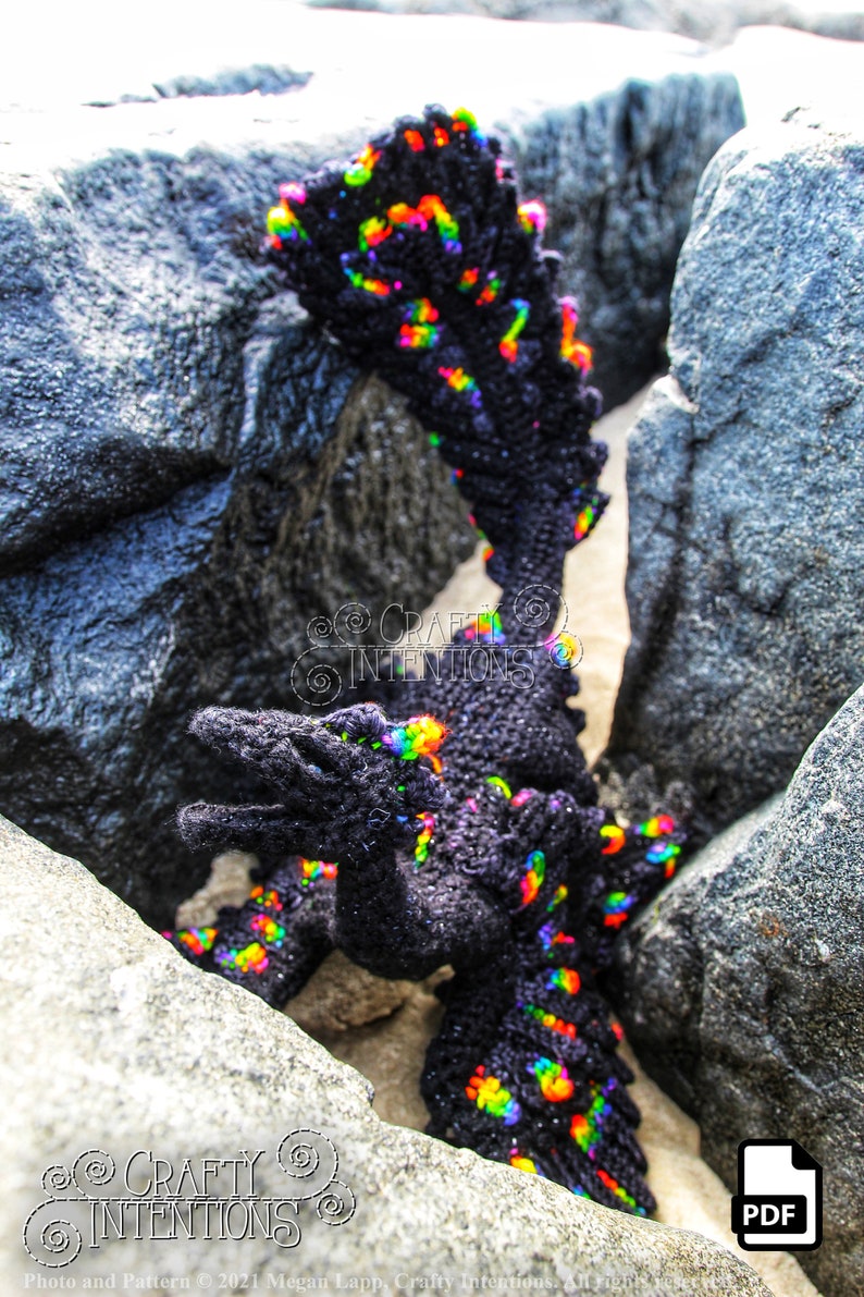 May include: A black crocheted dragon with rainbow coloured accents is posed in a rocky crevice. The dragon is standing on its hind legs with its front legs outstretched. The text "CRAFTY INTENTIONS" is visible in the image. A PDF icon is also visible in the image.