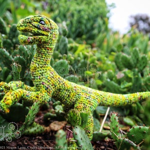 May include: A green crocheted dinosaur toy with a speckled pattern, posed on a bed of green cactus plants.