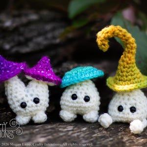 Puede incluir: Tres figuras de setas hechas a mano con ganchillo, con cuerpos blancos y sombreros coloridos en morado, turquesa y amarillo. Las setas tienen ojos negros y se exhiben sobre una superficie de madera natural. El texto "Crafty Intentions" es visible.