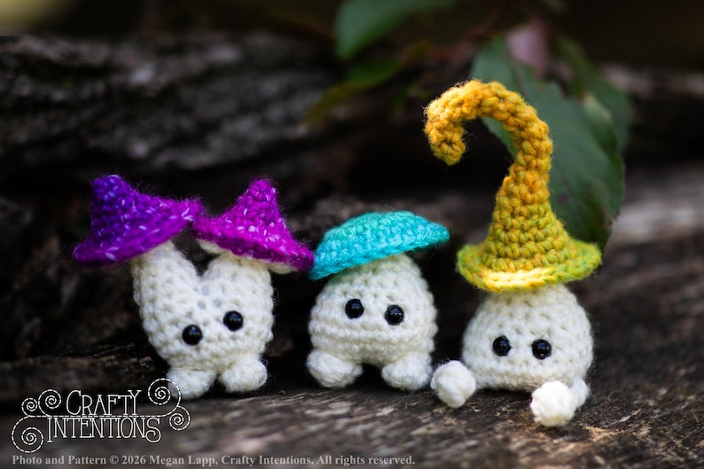 Puede incluir: Tres figuras de setas hechas a mano con ganchillo, con cuerpos blancos y sombreros coloridos en morado, turquesa y amarillo. Las setas tienen ojos negros y se exhiben sobre una superficie de madera natural. El texto "Crafty Intentions" es visible.
