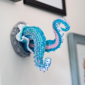 May include: A crocheted octopus wall hanging in shades of blue, pink, and white, mounted on a gray base. The tentacles curl upwards, showcasing a detailed, handmade design. The Crafty Intentions logo is visible.