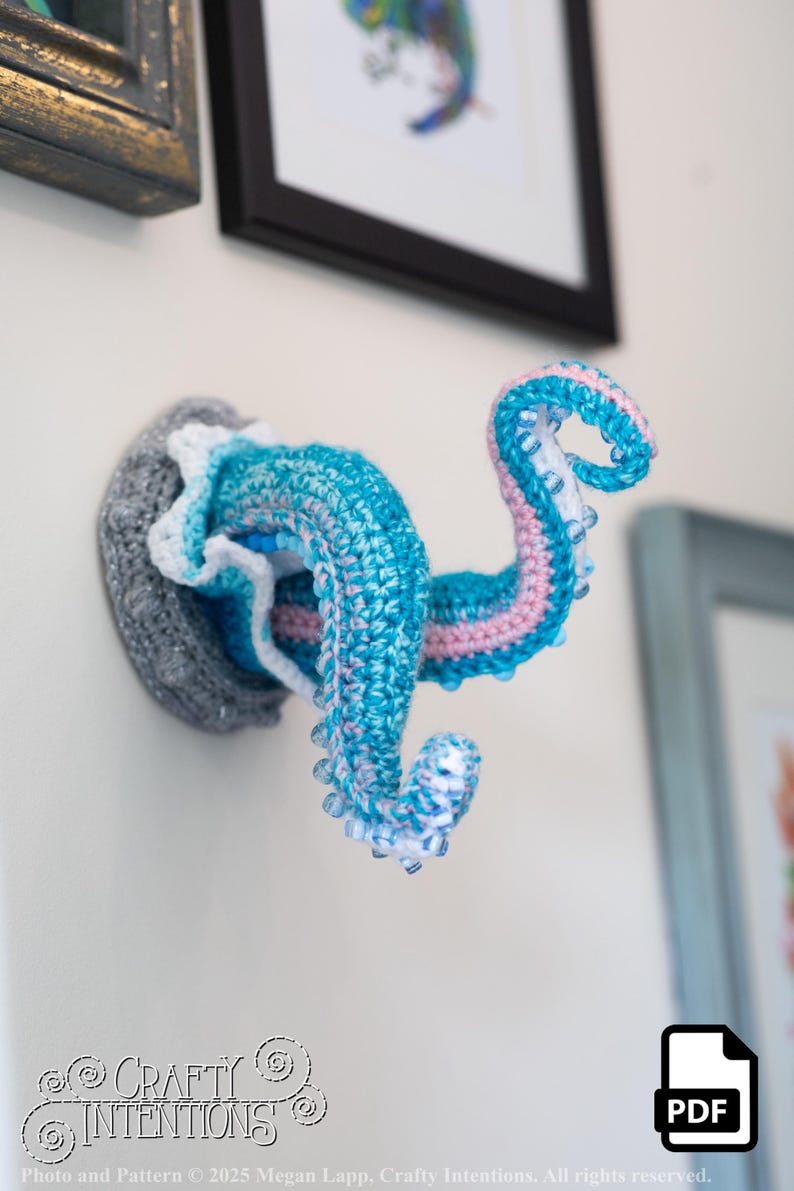 May include: A crocheted octopus wall hanging in shades of blue, pink, and white, mounted on a gray base. The tentacles curl upwards, showcasing a detailed, handmade design. The Crafty Intentions logo is visible.