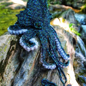 May include: A crocheted octopus amigurumi toy with a blue and green color scheme. The octopus is sitting on a piece of driftwood with a waterfall in the background. The toy is available as a PDF download.
