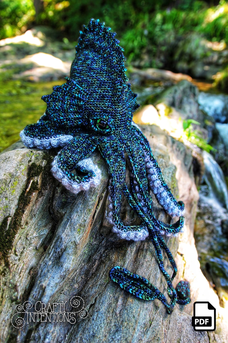 May include: A crocheted octopus amigurumi toy with a blue and green color scheme. The octopus is sitting on a piece of driftwood with a waterfall in the background. The toy is available as a PDF download.