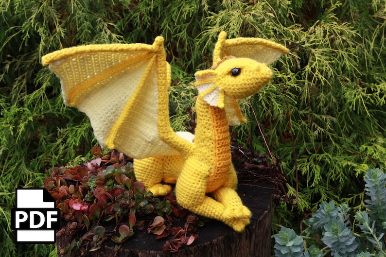 K&ouml;nnte beinhalten: Ein geh&auml;kelter gelber Drache mit langem Hals und gro&szlig;en Fl&uuml;geln. Der Drache sitzt auf einem Baumstumpf mit gr&uuml;nem Laub im Hintergrund.