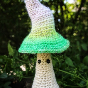 May include: A crocheted mushroom amigurumi toy with a white body, green and white hat, and black eyes. The toy is sitting on a wooden surface with green foliage in the background.