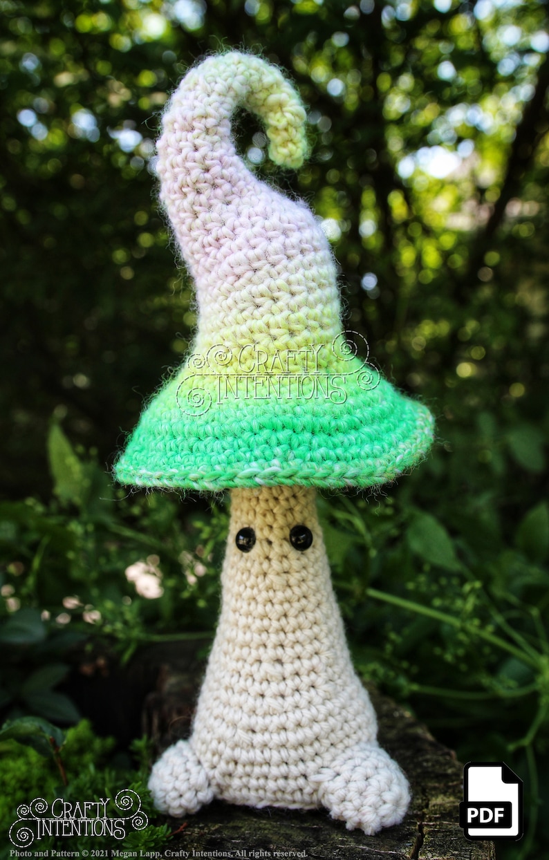 May include: A crocheted mushroom amigurumi toy with a white body, green and white hat, and black eyes. The toy is sitting on a wooden surface with green foliage in the background.