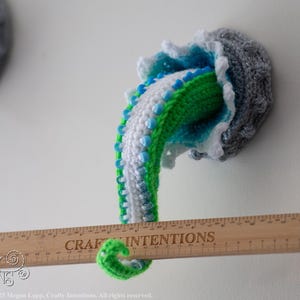 May include: Crocheted jellyfish wall art in shades of gray, blue, green, and white. The tentacles are green, white, and blue. A wooden ruler with the text "CRAFT INTENTIONS" is placed in front of the jellyfish. The jellyfish is mounted on a white wall.