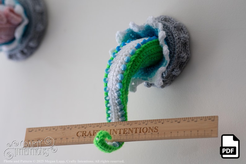 May include: Crocheted jellyfish wall art in shades of gray, blue, green, and white. The tentacles are green, white, and blue. A wooden ruler with the text "CRAFT INTENTIONS" is placed in front of the jellyfish. The jellyfish is mounted on a white wall.