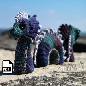 Może przedstawiać: Wężyk morski wykonany na szydełku w kolorze fioletowym, niebieskim i zielonym. Wąż ma długie, cienkie ciało z małą głową i dużym, okrągłym okiem. Wąż leży na piaszczystej plaży z niebieskim niebem w tle. Obraz zawiera również ikonę PDF z tekstem "PDF".