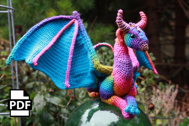 May include: A colorful crocheted dragon with a rainbow pattern. The dragon has a large, blue wing and is sitting on a green glass surface.