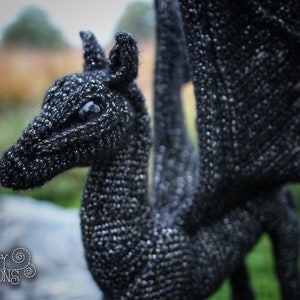 May include: A black crocheted dragon with a long tail and wings. The dragon has a detailed face with a large eye.