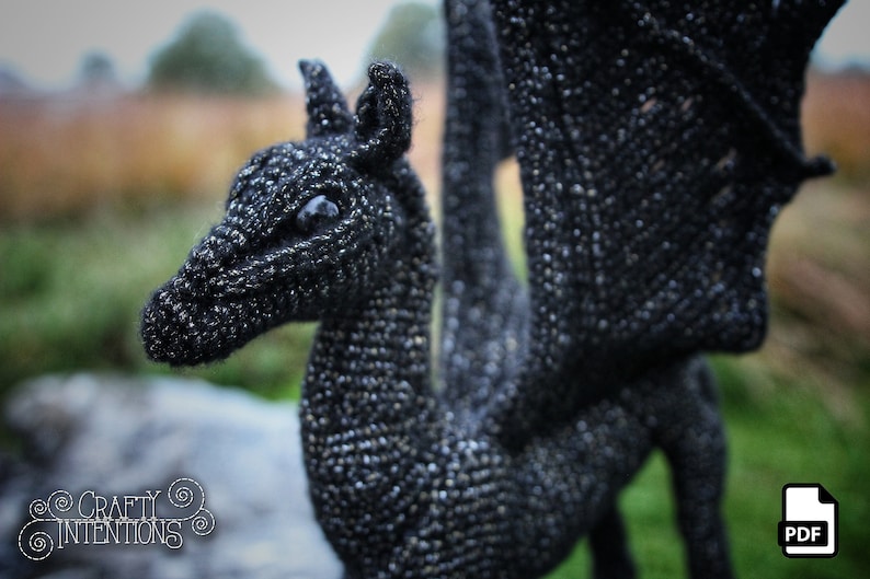 May include: A black crocheted dragon with a long tail and wings. The dragon has a detailed face with a large eye.