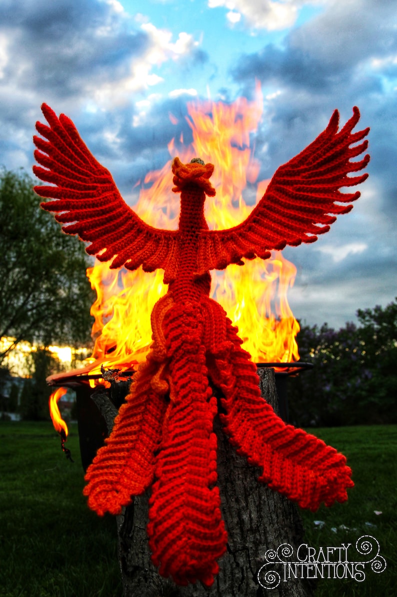 May include: A red crocheted phoenix rising from a fire pit. The phoenix has a long tail and outstretched wings. The fire is bright orange and yellow.
