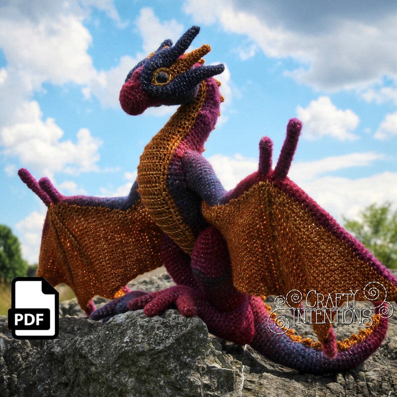 May include: A crocheted dragon with purple and orange wings, standing on a rock with a blue sky in the background. The dragon has a long tail and a small head with a pointed snout.