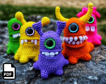 Può includere: Cinque mostri amigurumi all'uncinetto in diversi colori, tra cui verde, giallo, viola, arancione e rosa. Ogni mostro ha un solo occhio grande con una finitura scintillante e un ghigno pieno di denti. I mostri sono seduti su una superficie rocciosa grigia. Il testo "CRAFTY INTENTIONS" è visibile nell'angolo in basso a destra dell'immagine.