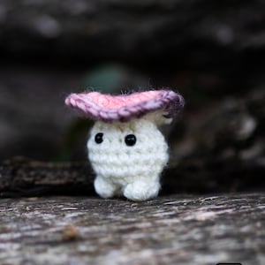 Puede incluir: Una peque&ntilde;a figura de hongo de ganchillo blanco con una gorra rosa y morada y ojos negros de bot&oacute;n. La figura est&aacute; sentada sobre una superficie de madera. La imagen incluye el texto "Crafty Intentions" y un icono PDF.