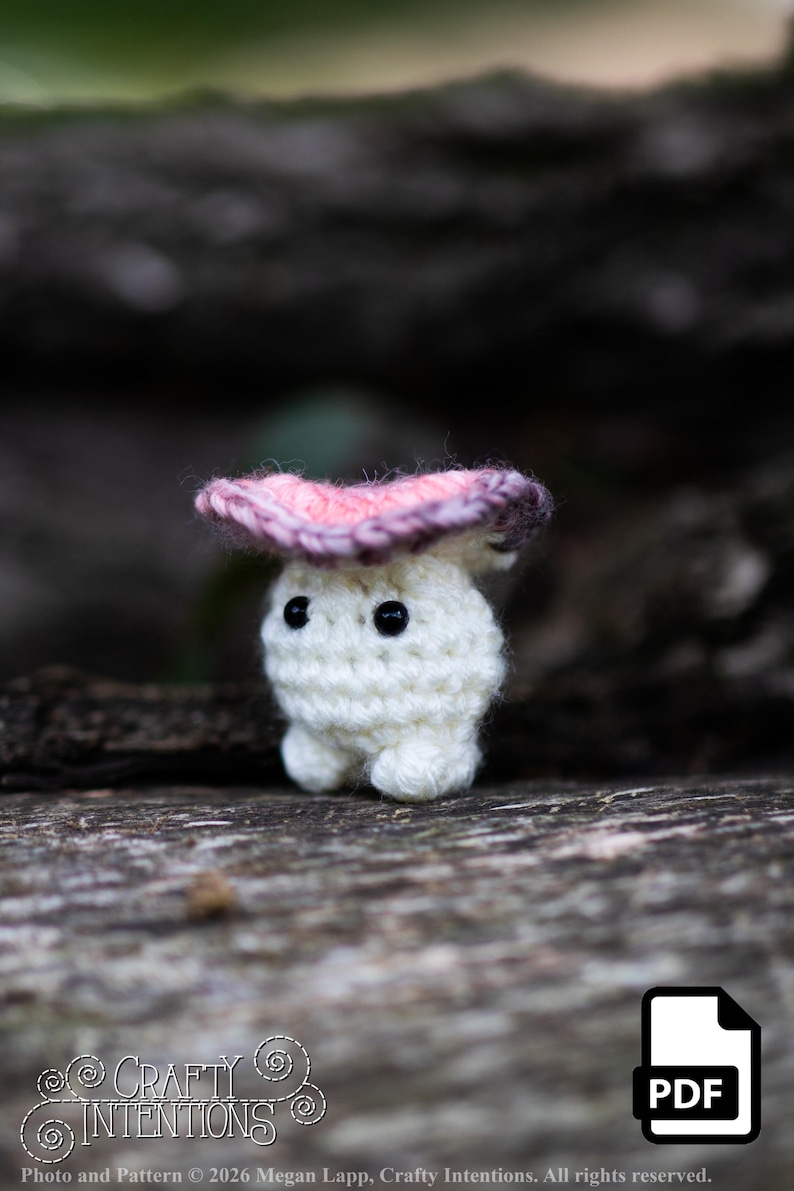 Puede incluir: Una peque&ntilde;a figura de hongo de ganchillo blanco con una gorra rosa y morada y ojos negros de bot&oacute;n. La figura est&aacute; sentada sobre una superficie de madera. La imagen incluye el texto "Crafty Intentions" y un icono PDF.
