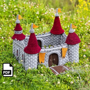 Könnte beinhalten: Eine gehäkelte Burg mit grauen Mauerwerk, burgunderroten Kegeldächern und orangefarbenen Flaggen. Die Burg hat einen braunen Bogeneingang und steht in einem Bett aus grünem Gras und weißen Blumen. Ein PDF-Symbol befindet sich in der unteren linken Ecke.