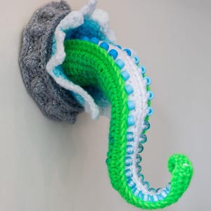 May include: A handmade, crocheted octopus tentacle wall hanging. The tentacle is green, white, and blue, with blue beads. The base is gray with silver accents. The design is whimsical and decorative. The image includes the text "Crafty Intentions" and a PDF icon.