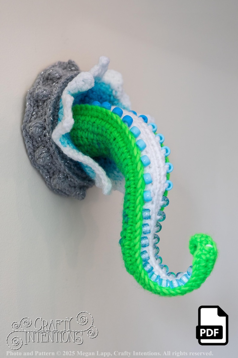 May include: A handmade, crocheted octopus tentacle wall hanging. The tentacle is green, white, and blue, with blue beads. The base is gray with silver accents. The design is whimsical and decorative. The image includes the text "Crafty Intentions" and a PDF icon.