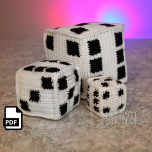 Puede incluir: Tres dados de crochet de diferentes tamaños, con un patrón de tablero de ajedrez en blanco y negro. Los dados están hechos de hilo blanco con cuadrados negros. El dado más grande está en la parte posterior, el dado mediano está en la parte delantera y el dado más pequeño está en la esquina delantera derecha. Los dados están sobre una superficie gris.