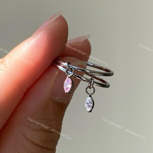 May include: A silver-toned double band ring featuring two dangling charms. One charm is a marquise-cut pink gemstone, and the other is a marquise-cut clear gemstone. The ring has a polished metallic finish and a simple, open-loop design.