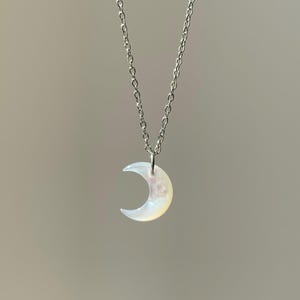 May include: A delicate silver necklace featuring a crescent moon pendant. The moon pendant has a pearlescent, iridescent finish, and hangs from a fine silver chain. The necklace is displayed against a neutral background.
