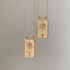 May include: Two gold-toned necklaces featuring rectangular pendants with a textured surface. Each pendant displays an embossed eye motif surrounded by radiating lines and small star shapes. The necklaces hang from delicate gold-toned metal chains.