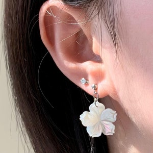 Może przedstawiać: Para kolczyków z białym kwiatem. Kolczyki są wykonane z perłowej masy perłowej i mają srebrne metalowe zapięcie.