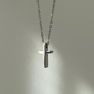 May include: A silver cross pendant necklace. The cross is small and suspended from a fine chain. The background is a soft, neutral colour, with subtle shadows adding depth. The necklace is simple and elegant.