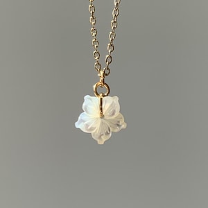Puede incluir: Un delicado collar dorado con un pequeño colgante de flor blanca de cinco pétalos. La flor tiene una apariencia translúcida y la cadena es de estilo eslabón fino. El fondo es un gris suave y neutro.