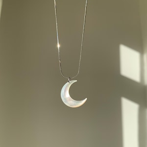 Può includere: Una collana d'argento con un ciondolo a forma di mezzaluna. Il ciondolo a forma di luna è bianco con riflessi iridescenti. La collana è posta su uno sfondo neutro, con ombre di luce solare.