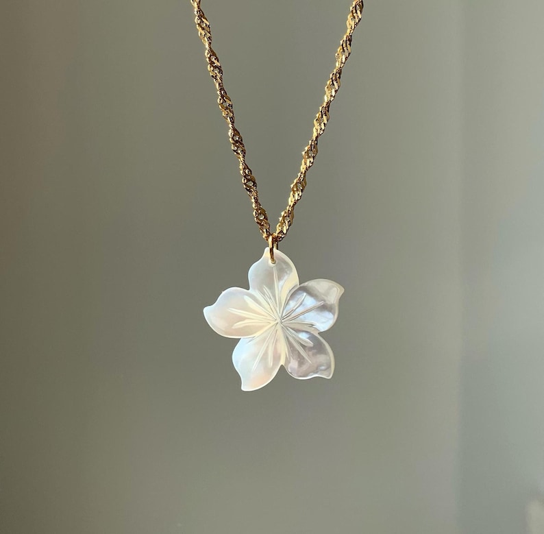 May include: A gold-colored chain necklace with a pendant shaped like a white flower. The flower pendant has five petals and a detailed, textured surface. The necklace is displayed against a neutral background.