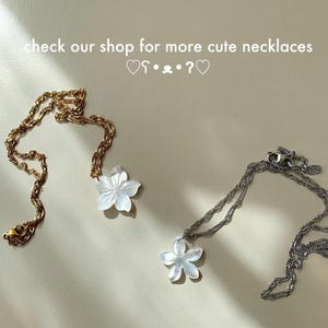 May include: Two necklaces with flower pendants. One necklace is gold-toned, the other is silver-toned. The flower pendants are white. The text "check our shop for more cute necklaces" is at the top of the image.