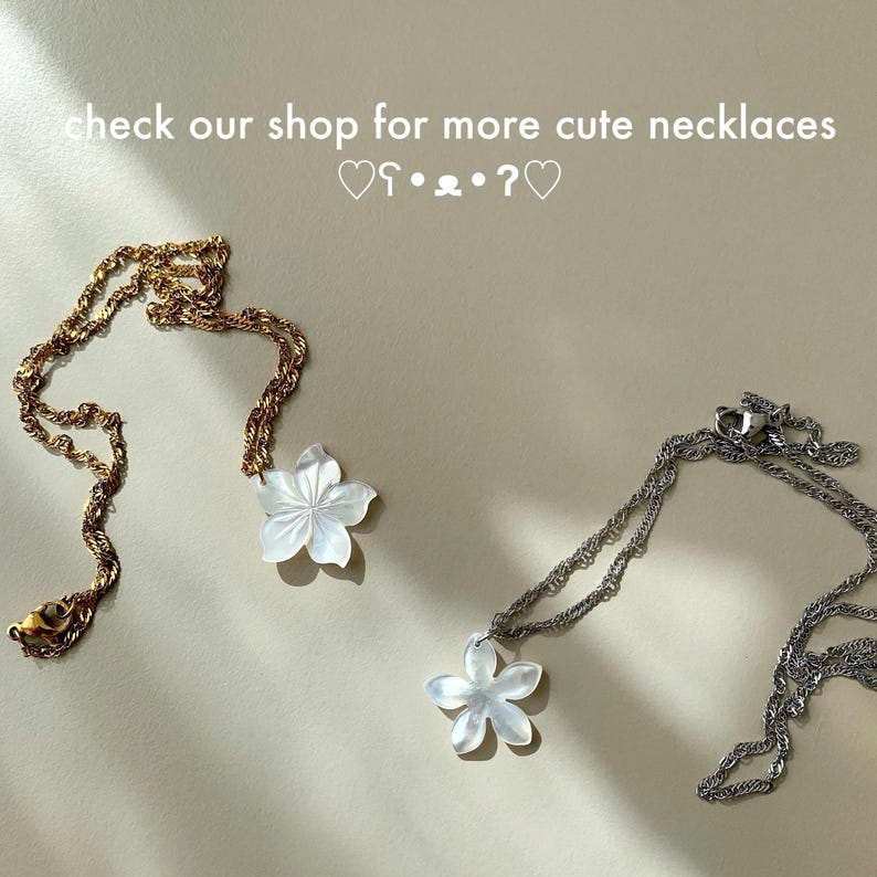 May include: Two necklaces with flower pendants. One necklace is gold-toned, the other is silver-toned. The flower pendants are white. The text "check our shop for more cute necklaces" is at the top of the image.