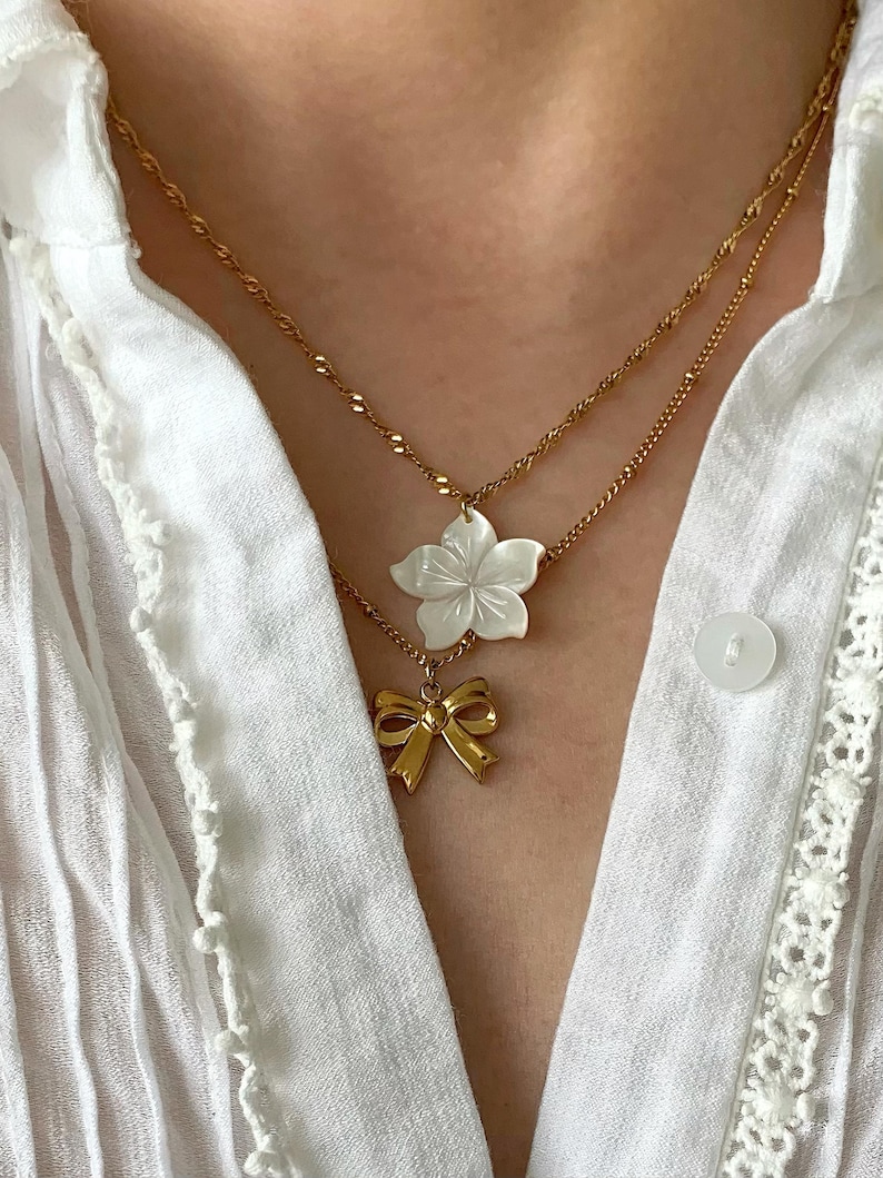 May include: Two gold necklaces with pendants. The top necklace features a white flower pendant, and the bottom necklace has a gold bow pendant. The necklaces are worn over a white blouse.
